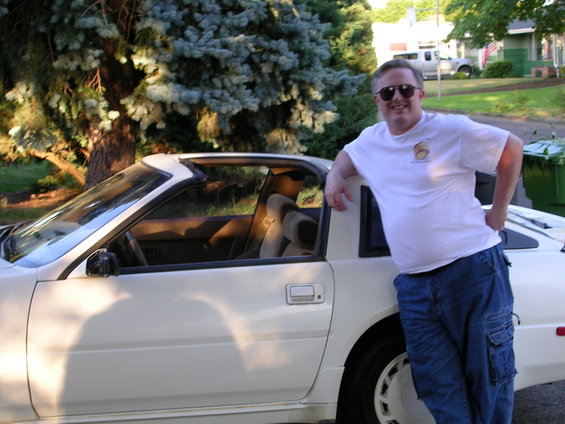 Man in sunglasses and white t-shirt leaning on a white 1980s sports car. Car has a t-top roof