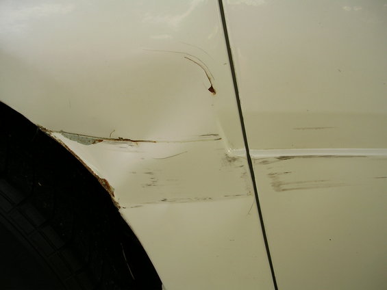 Close-up of a white car's side panel showing significant scratches, dents, and rust around the wheel well