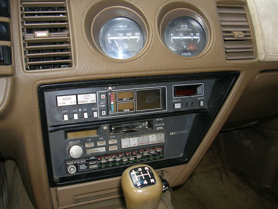 Car interior with tan dashboard, dual gauges, and a cassette radio. A manual gear shifter is visible