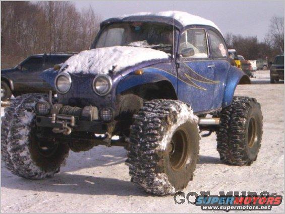 Modified Volkswagen Beetle with large off-road tires and snow on roof, parked in a snowy area with other vehicles in background