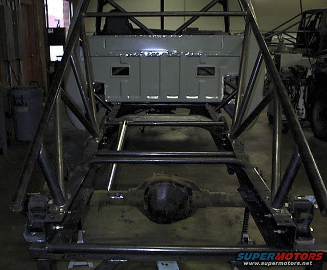 Truck chassis with custom roll cage and rear axle visible. Bed of truck is grey