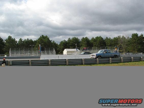 Blue pickup truck and black car on a race track with trees and bleachers in the background