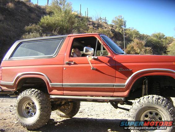 Side view of a red Ford Bronco with lifted suspension and large tires. A person is visible in the driver's seat
