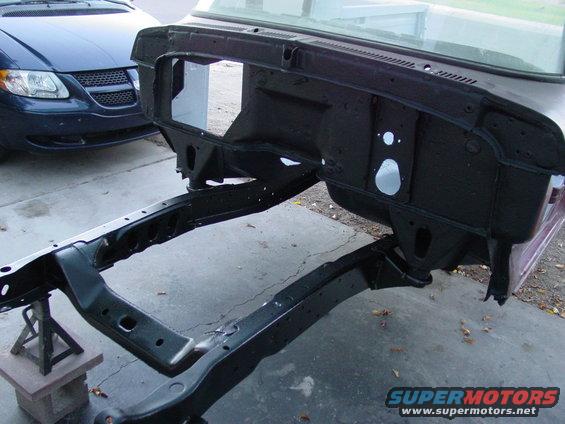 Vehicle frame and firewall assembly on jack stands. Blue minivan in background
