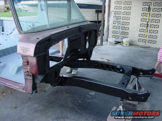 Vehicle frame and chassis on jack stands, with part of the body and windshield visible. Someone is standing behind it