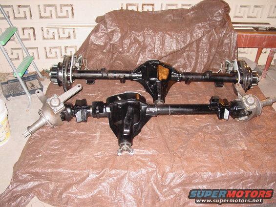 Two black Ford Bronco rear axles with disc brakes and brake lines, one above the other, on a brown tarp