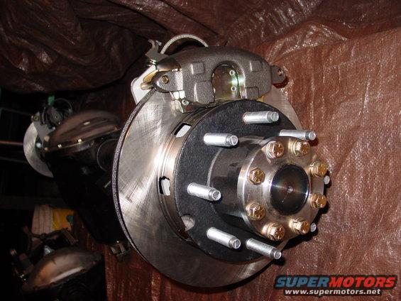 Close-up of a vehicle's rear brake rotor and caliper assembly, with a tarp in the background. Supermotors.net logo visible