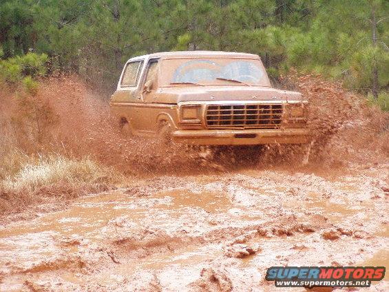 1978 Ford Bronco splashing through a muddy trail, trees in background