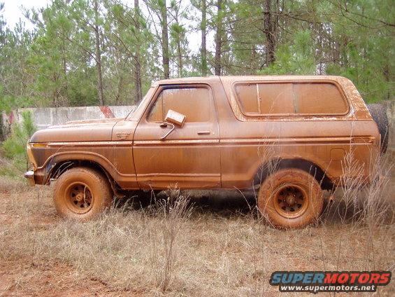 Side view of a 1978 Ford Bronco covered in mud, parked in a grassy, wooded area