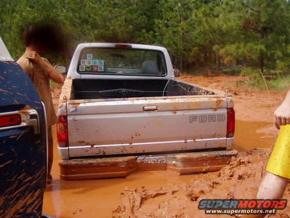Rear view of a silver Ford pickup truck stuck in mud, with a blue vehicle to its left