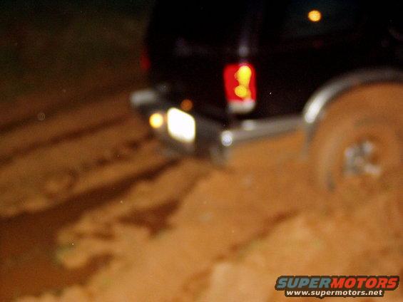 Rear of a dark-colored SUV driving through dirt, with taillights and backup lights illuminated