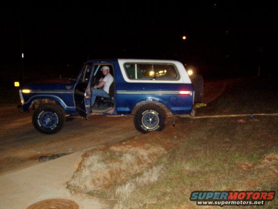 Blue and white 1978 Ford Bronco, driver's door open, parked on dirt and concrete at night