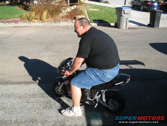 Man in black t-shirt and denim shorts rides a black mini motorcycle on a paved road