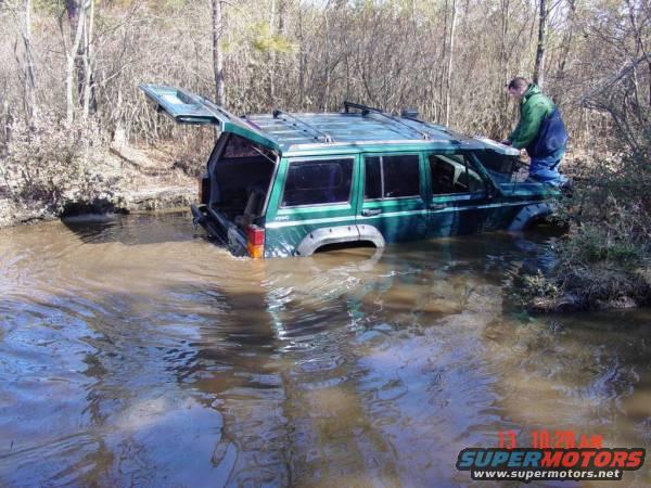 Green Jeep Cherokee stuck in muddy water, rear hatch open, person near driver's side