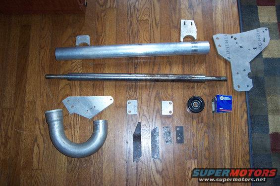 Various metal automotive parts including a large pipe, a shaft, brackets, a pulley, and a blue box on a wooden floor