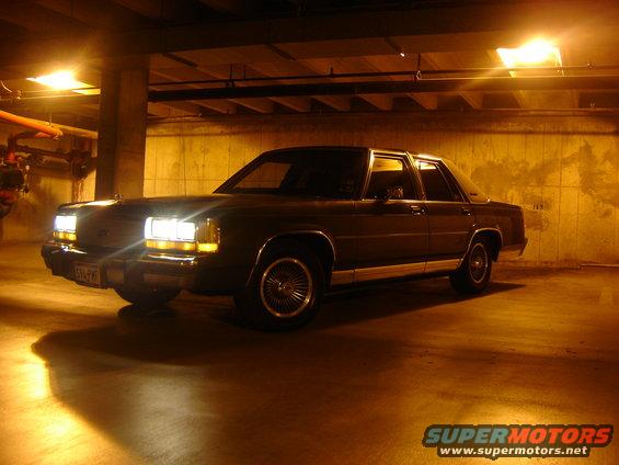 Dark sedan parked in a dimly lit garage, front quarter view, headlights on