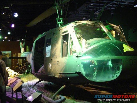 Green military helicopter with pilot mannequin inside, viewed from the front-right