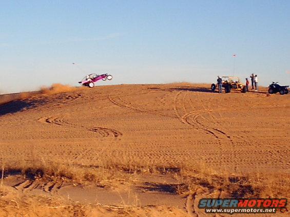 Pink dune buggy airborne over sand dune, other vehicles and people on ridge