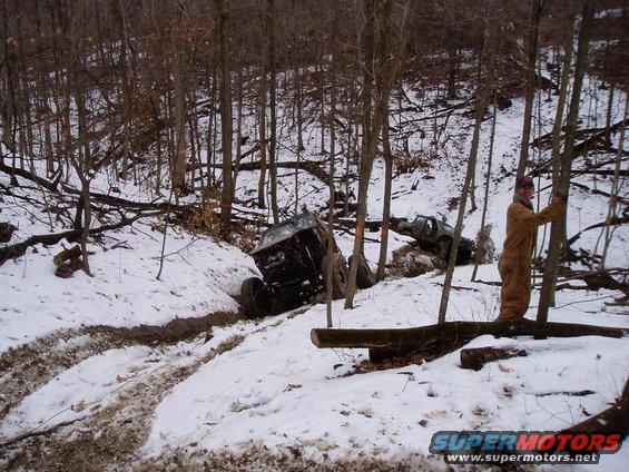 Two off-road vehicles stuck in a snowy ditch with trees and fallen logs around