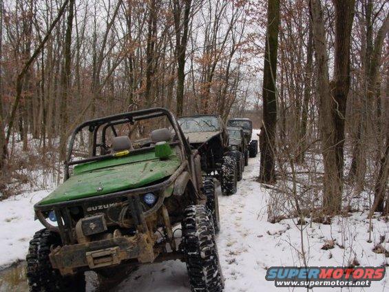 Green off-road vehicle with roll cage and large tires in snow, followed by other off-road vehicles