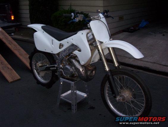 White dirt bike on a stand, front wheel visible, wooden ramps nearby