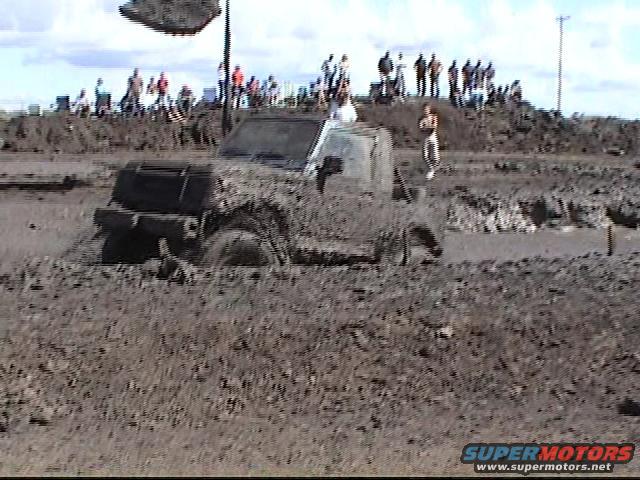 off-road vehicle stuck deep in mud pit with spectators on hill in background