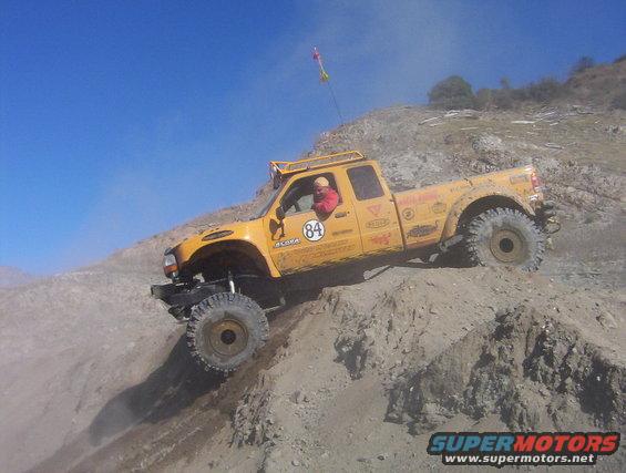 Yellow off-road pickup truck with large tires climbing a steep dirt hill. Driver is visible