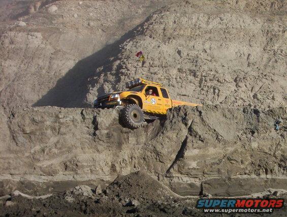 Yellow off-road truck climbing a steep dirt embankment, with a flag on its roof