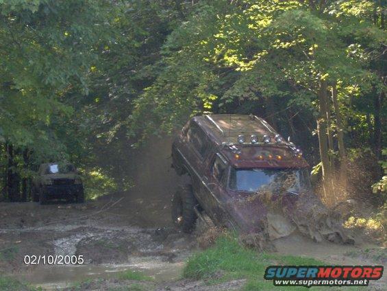 Two off-road vehicles navigating a muddy trail with trees in the background. One vehicle is partially submerged in mud