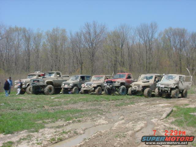 Several off-road vehicles lined up in a grassy, muddy area with trees in the background. A timestamp and website are visible in the lower right corner