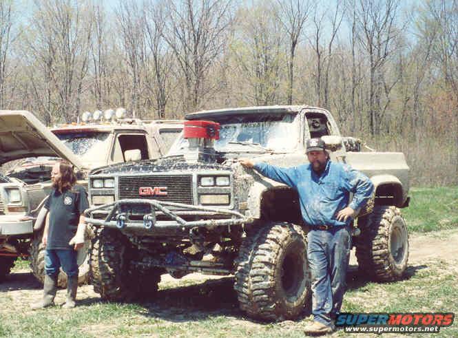 Mud-covered GMC pickup truck with large tires and exposed engine, a man leans on it