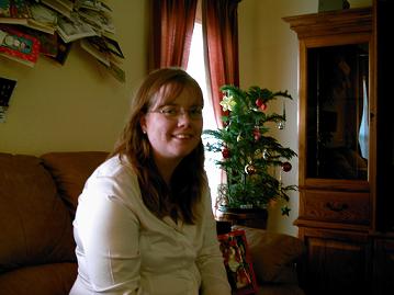 Woman wearing glasses and a white shirt sits on a brown couch next to a small Christmas tree