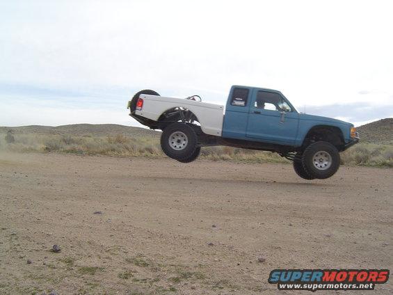 Blue and white pickup truck mid-air over a dirt road, with a spare tire mounted on the back