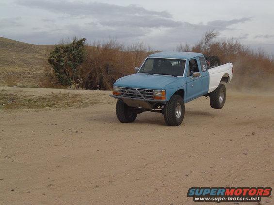 Blue and white pickup truck with off-road modifications airborne on a dirt track