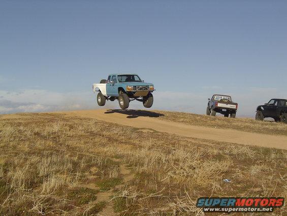 Blue and white pickup truck mid-air over a dirt hill, with two other trucks behind it