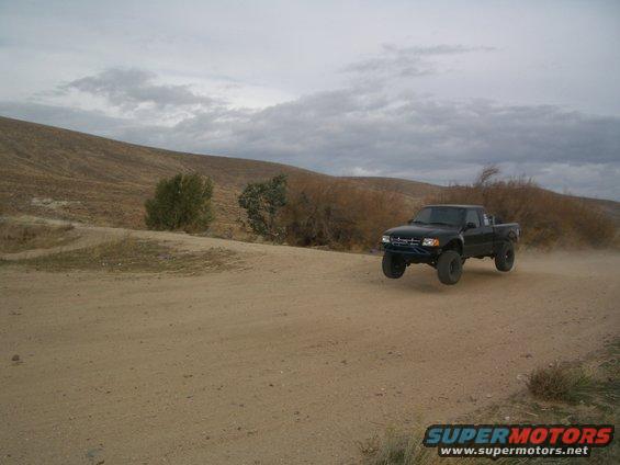 black pickup truck mid-air on a dirt road, kicking up dust. Hills and cloudy sky in background