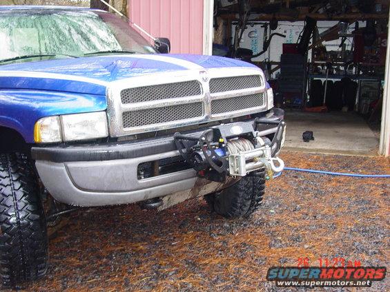 Front view of a blue Dodge pickup truck with a winch mounted on the bumper. The truck has a chrome grille and white racing stripes on the hood