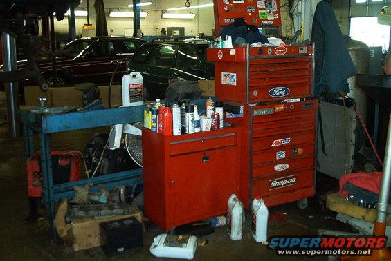 Two cars in a garage, one maroon sedan and one dark green SUV, with toolboxes and automotive fluids