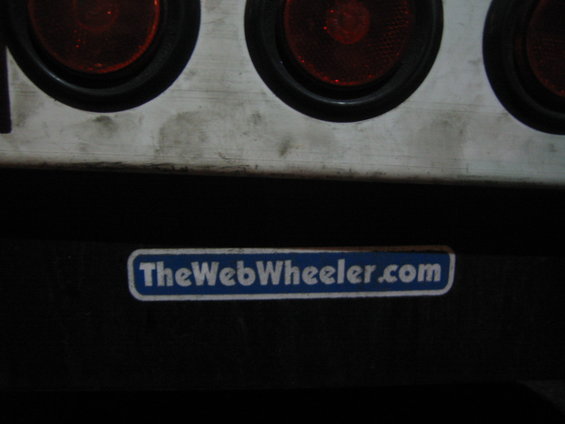 Close-up of dirty white panel with four red tail lights above a blue sticker reading "TheWebWheeler.com"