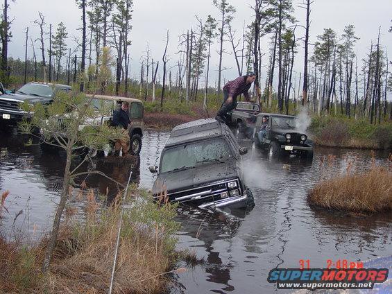 Black van partially submerged in water, front view. Water splashes around it. Other off-road vehicles visible