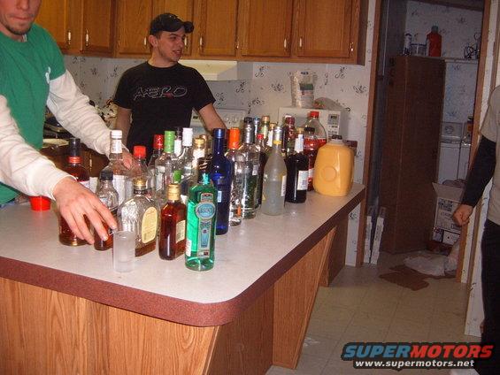 People gathered around a kitchen counter loaded with various bottles of alcohol and a large orange jug