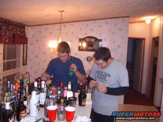 Two men pour drinks from bottles on a table laden with alcohol and red cups