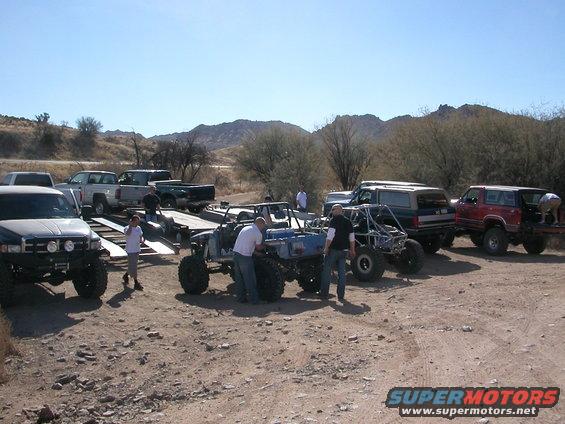 Several off-road vehicles and trucks parked in a dirt lot with mountains in the background