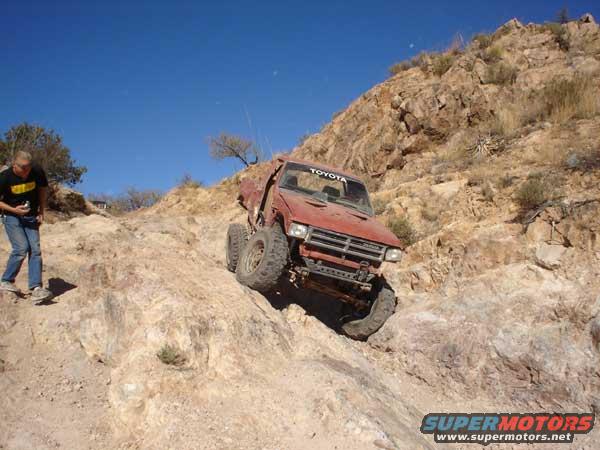 Red Toyota truck with off-road modifications climbing a steep, rocky incline under a clear blue sky