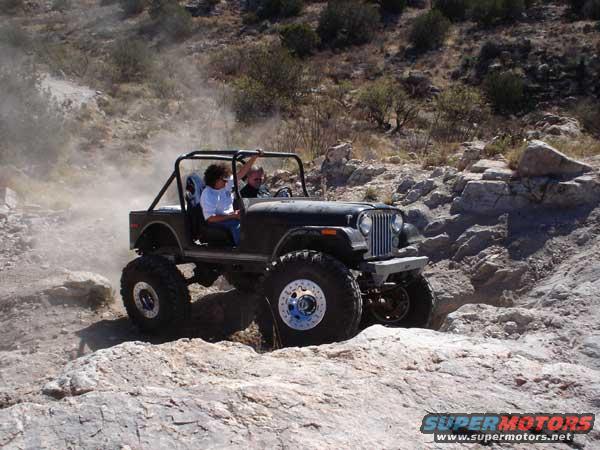 Jeep CJ-7 driving on rocky terrain, kicking up dust. Two people are inside