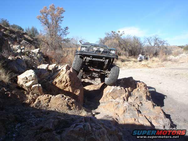 1989 Ford Bronco climbing over large rocks on a dirt trail, another vehicle visible in background