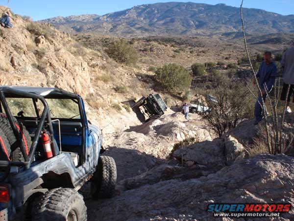 Several off-road vehicles navigate a steep, rocky trail in a desert landscape. One vehicle is in the foreground, facing away