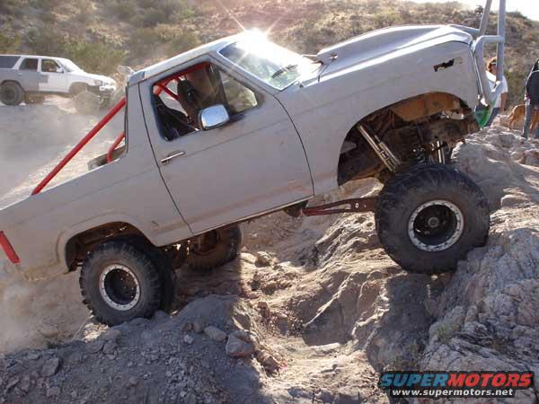 Gray off-road truck with red roll cage climbing rocks, another white SUV in background