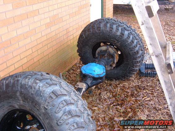 Two large off-road tires and a vehicle axle with a blue differential cover sit on the ground