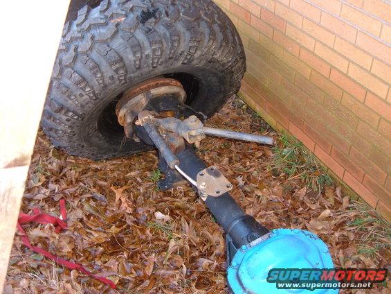 Vehicle axle with large tire, sitting on the ground near a brick wall